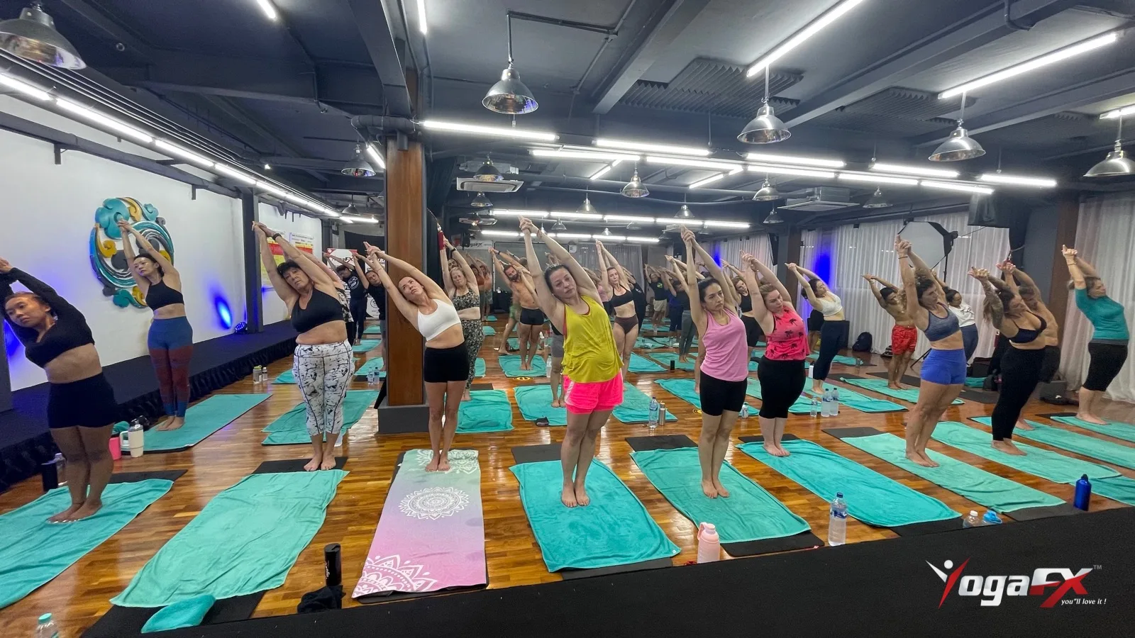 Students reaching overhead in a held half-moon side stretch while the teacher guides the class from the stage